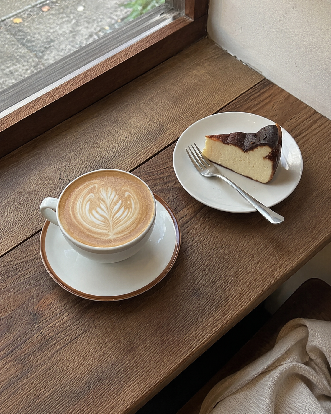 窓辺の木のベンチに置かれた、ラテアートのカフェラテとバスクチーズケーキ。やわらかな自然光